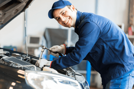 mechanic in a blue overall lining over open hood of a car with a wrench